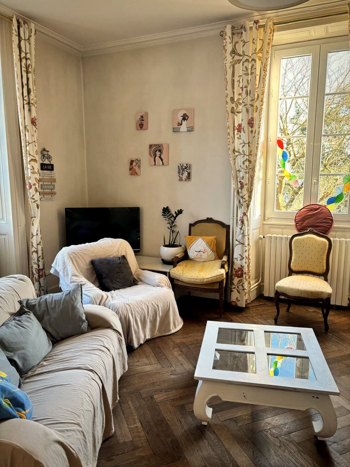 Le salon de Laval, avec ses canapés clairs, ses fauteuils et ses grandes fenêtres à rideaux fleuris.
