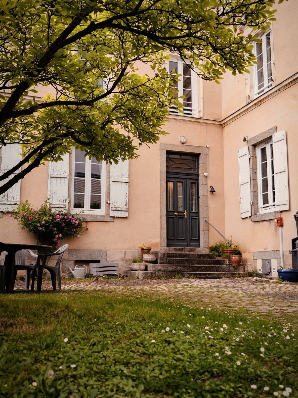 La façade de la maison de Laval, une maison ancienne aux volets clairs, avec un arbre et un rosier dans la cour.
